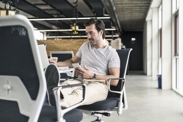 Junger Geschäftsmann arbeitet entspannt in einem modernen Büro, mit Smartphone und Laptop - UUF17815