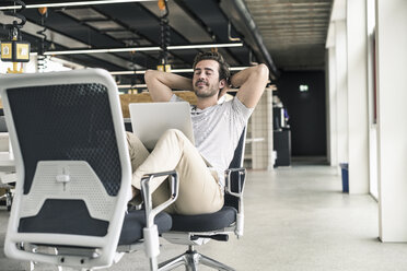 Junger Geschäftsmann arbeitet entspannt in einem modernen Büro, mit Laptop auf dem Schoß - UUF17818