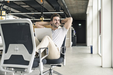 Junger Geschäftsmann arbeitet entspannt in einem modernen Büro, mit Laptop auf dem Schoß - UUF17819