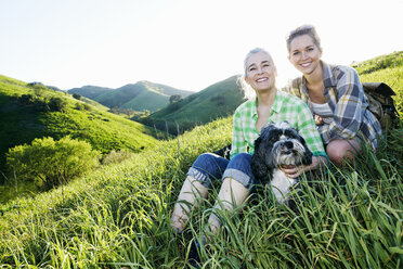 Caucasian mother and daughter petting dog on grassy hillside - BLEF06590