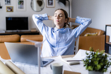 Woman in office leaning back with plan, laptop and wind turbine model on table - GIOF06434