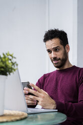 Junger Mann sitzt zu Hause am Tisch und benutzt Laptop und Mobiltelefon - GIOF06461