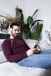 Lächelnder Mann sitzt mit Handy und Kopfhörern auf der Couch - GIOF06479