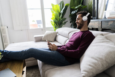 Laughing man sitting on couch with cell phone and headphones - GIOF06480