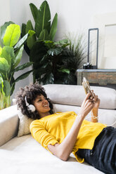 Smiling woman lying on couch with cell phone and headphones - GIOF06487