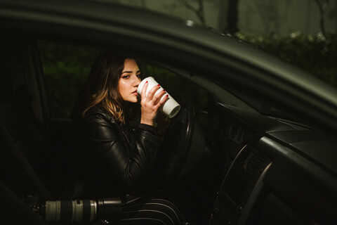 Junge Frau mit Einwegbecher und Kamera in einem Auto bei Nacht, lizenzfreies Stockfoto