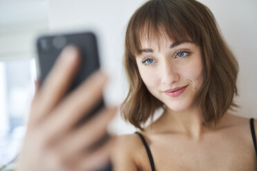 Porträt einer lächelnden jungen Frau, die zu Hause ein Selfie mit ihrem Smartphone macht - PNEF01544