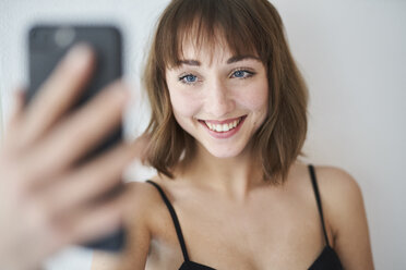 Porträt einer lächelnden jungen Frau, die zu Hause ein Selfie mit ihrem Smartphone macht - PNEF01545