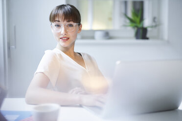 Portrait of young businesswoman with laptop sitting at desk in an office - PNEF01548