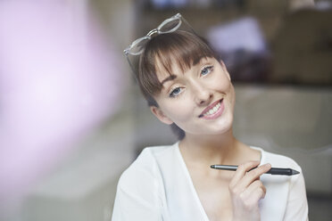 Portrait of smiling young businesswoman with glasses and pencil behind glass pane - PNEF01554