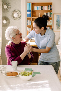Lächelnde ältere Frau im Ruhestand, die die Hände einer jungen Freiwilligen hält, während sie im Pflegeheim spricht - MASF12362