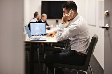 Young businessman talking on phone while using laptop at conference table in board room - MASF12391