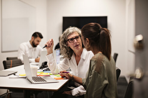 Geschäftsfrauen diskutieren über einem Laptop, während ein männlicher Kollege im Hintergrund arbeitet - MASF12393