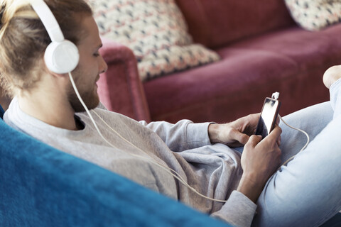 Junger Mann sitzt mit Handy und Kopfhörern auf der Couch, lizenzfreies Stockfoto