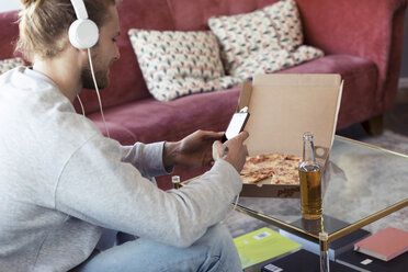 Junger Mann auf der Couch mit Handy und Kopfhörern beim Pizzaessen - JSRF00181