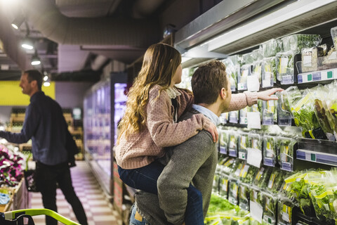 Seitenansicht einer Tochter, die ein Gemüsepaket aufhebt, während sie von ihrem Vater im Laden huckepack genommen wird, lizenzfreies Stockfoto