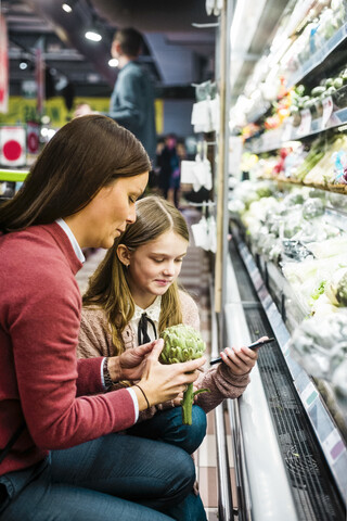 Mutter hält Artischocke, während ihre Tochter im Supermarkt ein Mobiltelefon benutzt, lizenzfreies Stockfoto