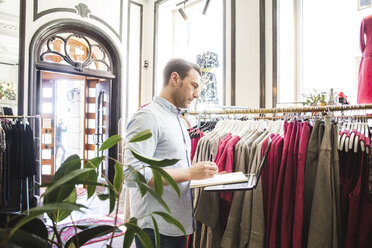 Seitenansicht einer Verkäuferin mit Blick auf einen Kleiderständer, die ein Buch und einen Laptop in einer Boutique hält - MASF12444