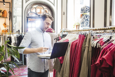 Selbstbewusster männlicher Unternehmer, der einen Laptop benutzt, während er an einem Kleiderständer in einer Boutique steht - MASF12447