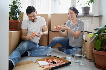 Smiling couple eating pizza while sitting against cardboard boxes in new house - MASF12506