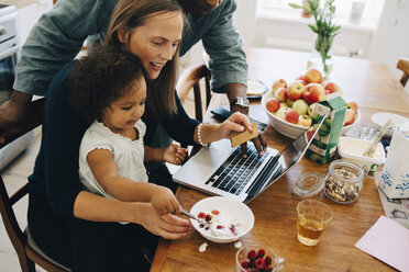 Parents shopping online on laptop while looking at daughter having breakfast in dining room - MASF12542