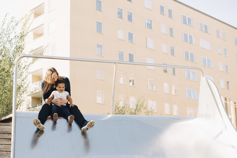 Mother playing with daughter on slide at park in city stock photo