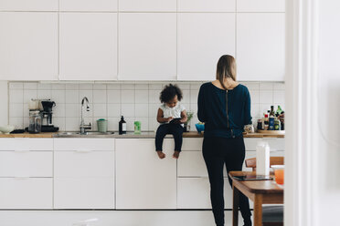 Mädchen in voller Länge, das ein Smartphone benutzt, während die Mutter in der Küche zu Hause arbeitet - MASF12568