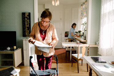 Senior woman reading newspaper while caregiver working in kitchen at nursing home - MASF12580