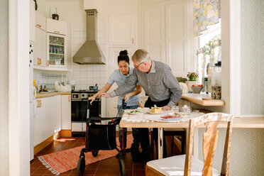 Young female volunteer assisting retired elderly man with rollator in kitchen at nursing home - MASF12589