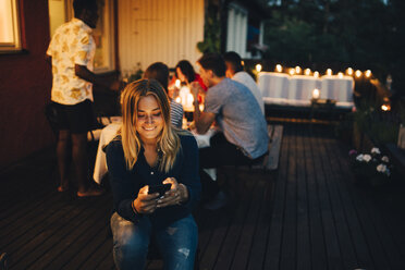 Lächelnde junge Frau, die ein Smartphone benutzt, während Freunde im Hintergrund auf einer Dinnerparty sind - MASF12606
