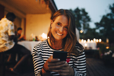 Porträt einer lächelnden Frau, die ein Smartphone in der Hand hält, während im Hintergrund Freunde auf einer Dinnerparty sitzen - MASF12608