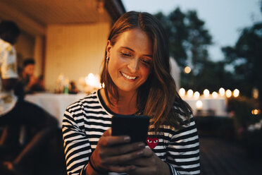 Smiling young woman using smart phone while friends in background during dinner party - MASF12609