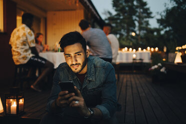 Portrait of smiling man holding smart phone while friends in background during dinner party - MASF12614