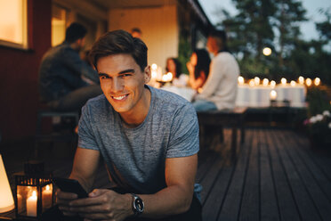 Porträt eines lächelnden jungen Mannes, der sein Smartphone in der Hand hält, während im Hintergrund Freunde auf einer Dinnerparty sitzen - MASF12615