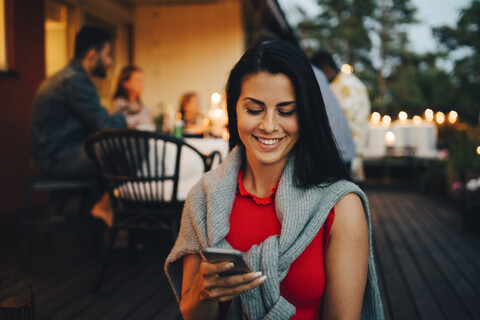 Lächelnde Frau im mittleren Erwachsenenalter, die ein Mobiltelefon benutzt, während Freunde im Hintergrund auf einer Dinnerparty sind, lizenzfreies Stockfoto