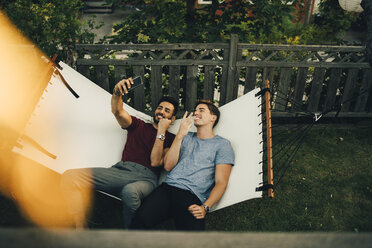 High angle view of smiling friends taking selfie on smart phone while resting in hammock at yard - MASF12651