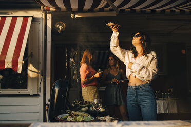 Frau fotografiert mit ihrem Smartphone Essen auf dem Grill, während Freunde im Hintergrund stehen - MASF12672