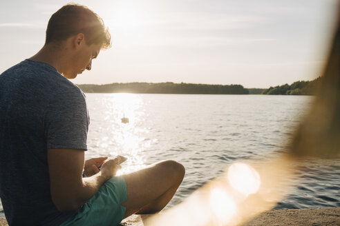 Junger Mann, der ein Mobiltelefon benutzt, während er im Sommer am Steg am See sitzt - MASF12702