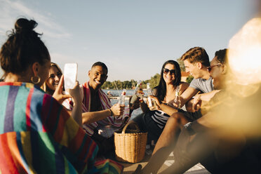 Woman photographing cheerful friends toasting drinks at jetty in summer - MASF12703