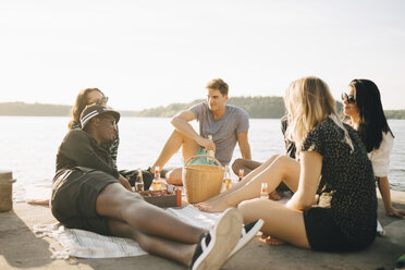 Multi-ethnic friends enjoying food and drink while sitting on jetty at lake against sky - MASF12709