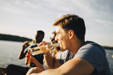 Man enjoying drink with friends while sitting on jetty against sky in summer - MASF12712