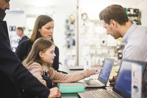 Niedliches Mädchen zeigt auf den Bildschirm eines Laptops auf einen Verkäufer, während sie mit ihrer Familie in einem Computerladen steht - MASF12742