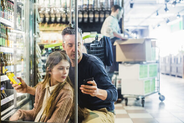 Vater und Tochter schauen auf ihr Mobiltelefon, während sie in einem Geschäft am Kühlschrank kauern - MASF12768