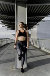 Sportliche Frau mit Kopfhörern, die auf einer Brücke steht, Musik hört und ein Smartphone benutzt - ERRF01462