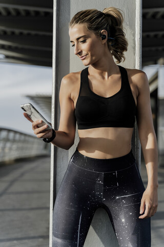 Sportliche Frau mit Kopfhörern, die auf einer Brücke steht, Musik hört und ein Smartphone benutzt, lizenzfreies Stockfoto