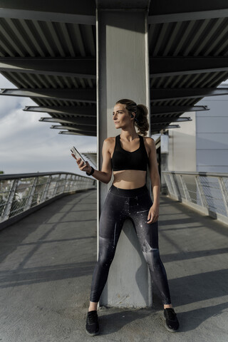 Sportliche Frau mit Kopfhörern, die auf einer Brücke steht, Musik hört und ein Smartphone benutzt, lizenzfreies Stockfoto