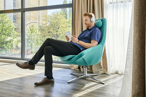 Junger Mann sitzt im Sessel, hört Musik und benutzt ein digitales Tablet, lizenzfreies Stockfoto