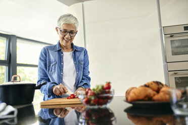 Senior woman standing in kitchen, chopping strawberries - ZEDF02315