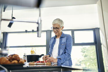 Senior woman standing in kitchen, chopping strawberries - ZEDF02318