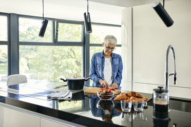 Senior woman standing in kitchen, chopping strawberries - ZEDF02319